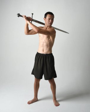Yakışıklı Asyalı erkek modelin tam boy portresi, spor şortu ve gömleksiz. Beyaz stüdyo arka planında izole edilmiş, savaşçı eğitimi sırasında kılıçlarını tutuyor..
