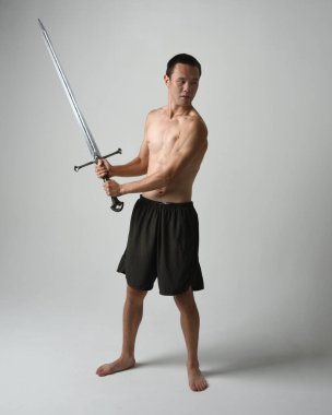 Yakışıklı Asyalı erkek modelin tam boy portresi, spor şortu ve gömleksiz. Beyaz stüdyo arka planında izole edilmiş, savaşçı eğitimi sırasında kılıçlarını tutuyor..