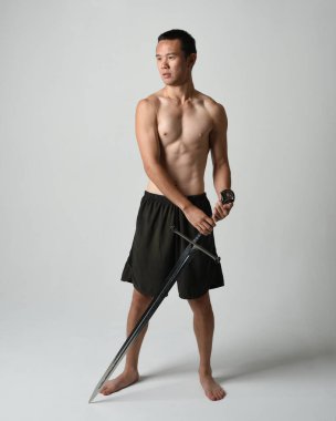 Yakışıklı Asyalı erkek modelin tam boy portresi, spor şortu ve gömleksiz. Beyaz stüdyo arka planında izole edilmiş, savaşçı eğitimi sırasında kılıçlarını tutuyor..