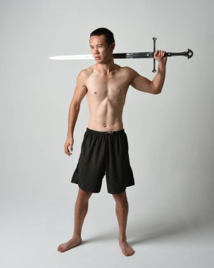 Yakışıklı Asyalı erkek modelin tam boy portresi, spor şortu ve gömleksiz. Beyaz stüdyo arka planında izole edilmiş, savaşçı eğitimi sırasında kılıçlarını tutuyor..
