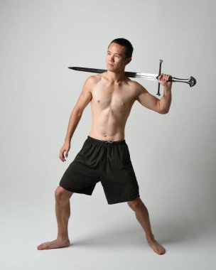 Yakışıklı Asyalı erkek modelin tam boy portresi, spor şortu ve gömleksiz. Beyaz stüdyo arka planında izole edilmiş, savaşçı eğitimi sırasında kılıçlarını tutuyor..