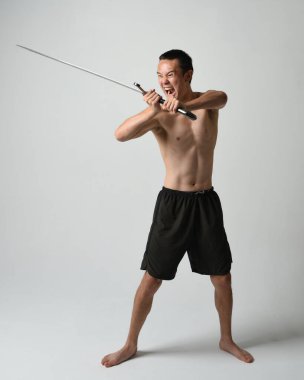 Yakışıklı Asyalı erkek modelin tam boy portresi, spor şortu ve gömleksiz. Beyaz stüdyo arka planında izole edilmiş, savaşçı eğitimi sırasında kılıçlarını tutuyor..