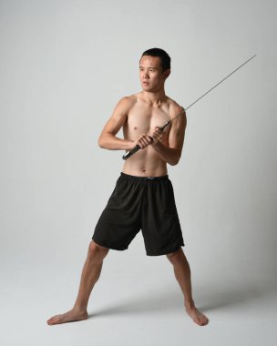Yakışıklı Asyalı erkek modelin tam boy portresi, spor şortu ve gömleksiz. Beyaz stüdyo arka planında izole edilmiş, savaşçı eğitimi sırasında kılıçlarını tutuyor..