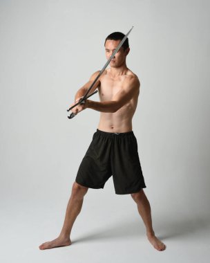 Yakışıklı Asyalı erkek modelin tam boy portresi, spor şortu ve gömleksiz. Beyaz stüdyo arka planında izole edilmiş, savaşçı eğitimi sırasında kılıçlarını tutuyor..