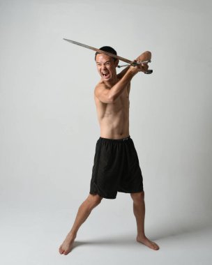 Yakışıklı Asyalı erkek modelin tam boy portresi, spor şortu ve gömleksiz. Beyaz stüdyo arka planında izole edilmiş, savaşçı eğitimi sırasında kılıçlarını tutuyor..
