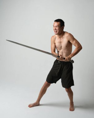 Yakışıklı Asyalı erkek modelin tam boy portresi, spor şortu ve gömleksiz. Beyaz stüdyo arka planında izole edilmiş, savaşçı eğitimi sırasında kılıçlarını tutuyor..