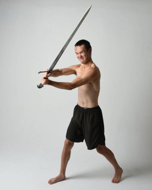 Yakışıklı Asyalı erkek modelin tam boy portresi, spor şortu ve gömleksiz. Beyaz stüdyo arka planında izole edilmiş, savaşçı eğitimi sırasında kılıçlarını tutuyor..