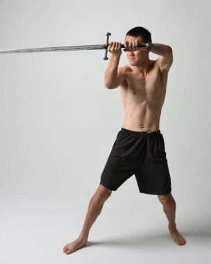 Yakışıklı Asyalı erkek modelin tam boy portresi, spor şortu ve gömleksiz. Beyaz stüdyo arka planında izole edilmiş, savaşçı eğitimi sırasında kılıçlarını tutuyor..