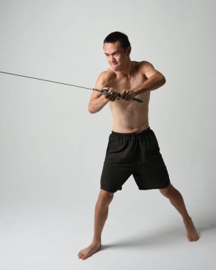 Yakışıklı Asyalı erkek modelin tam boy portresi, spor şortu ve gömleksiz. Beyaz stüdyo arka planında izole edilmiş, savaşçı eğitimi sırasında kılıçlarını tutuyor..