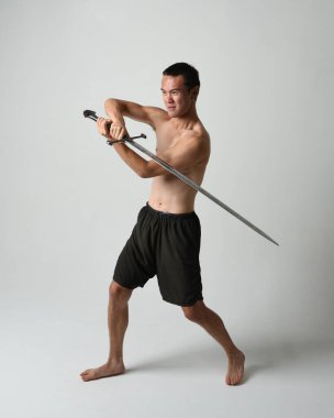 Yakışıklı Asyalı erkek modelin tam boy portresi, spor şortu ve gömleksiz. Beyaz stüdyo arka planında izole edilmiş, savaşçı eğitimi sırasında kılıçlarını tutuyor..