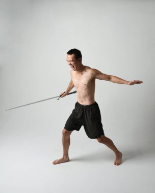 Yakışıklı Asyalı erkek modelin tam boy portresi, spor şortu ve gömleksiz. Beyaz stüdyo arka planında izole edilmiş, savaşçı eğitimi sırasında kılıçlarını tutuyor..