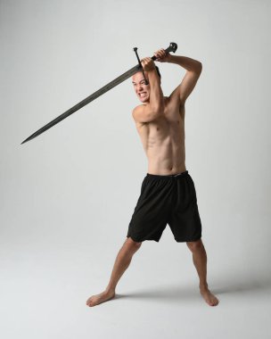 Yakışıklı Asyalı erkek modelin tam boy portresi, spor şortu ve gömleksiz. Beyaz stüdyo arka planında izole edilmiş, savaşçı eğitimi sırasında kılıçlarını tutuyor..