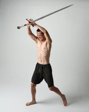 Yakışıklı Asyalı erkek modelin tam boy portresi, spor şortu ve gömleksiz. Beyaz stüdyo arka planında izole edilmiş, savaşçı eğitimi sırasında kılıçlarını tutuyor..