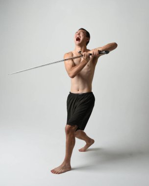 Yakışıklı Asyalı erkek modelin tam boy portresi, spor şortu ve gömleksiz. Beyaz stüdyo arka planında izole edilmiş, savaşçı eğitimi sırasında kılıçlarını tutuyor..