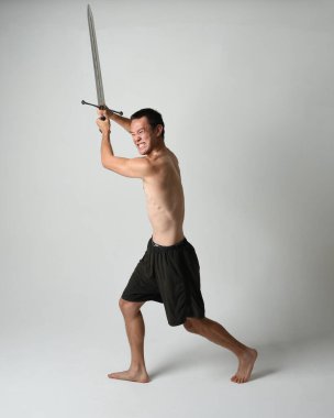 Yakışıklı Asyalı erkek modelin tam boy portresi, spor şortu ve gömleksiz. Beyaz stüdyo arka planında izole edilmiş, savaşçı eğitimi sırasında kılıçlarını tutuyor..