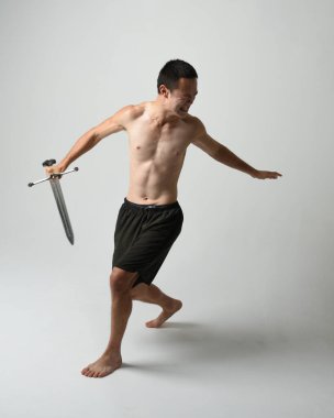 Yakışıklı Asyalı erkek modelin tam boy portresi, spor şortu ve gömleksiz. Beyaz stüdyo arka planında izole edilmiş, savaşçı eğitimi sırasında kılıçlarını tutuyor..
