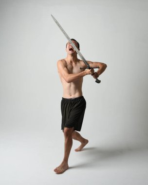Yakışıklı Asyalı erkek modelin tam boy portresi, spor şortu ve gömleksiz. Beyaz stüdyo arka planında izole edilmiş, savaşçı eğitimi sırasında kılıçlarını tutuyor..