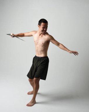 Yakışıklı Asyalı erkek modelin tam boy portresi, spor şortu ve gömleksiz. Beyaz stüdyo arka planında izole edilmiş, savaşçı eğitimi sırasında kılıçlarını tutuyor..