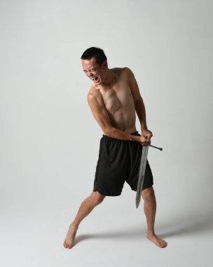 Yakışıklı Asyalı erkek modelin tam boy portresi, spor şortu ve gömleksiz. Beyaz stüdyo arka planında izole edilmiş, savaşçı eğitimi sırasında kılıçlarını tutuyor..