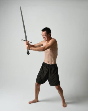 Yakışıklı Asyalı erkek modelin tam boy portresi, spor şortu ve gömleksiz. Beyaz stüdyo arka planında izole edilmiş, savaşçı eğitimi sırasında kılıçlarını tutuyor..