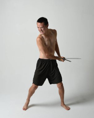 Yakışıklı Asyalı erkek modelin tam boy portresi, spor şortu ve gömleksiz. Beyaz stüdyo arka planında izole edilmiş, savaşçı eğitimi sırasında kılıçlarını tutuyor..