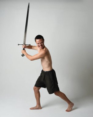 Yakışıklı Asyalı erkek modelin tam boy portresi, spor şortu ve gömleksiz. Beyaz stüdyo arka planında izole edilmiş, savaşçı eğitimi sırasında kılıçlarını tutuyor..