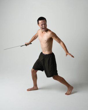 Yakışıklı Asyalı erkek modelin tam boy portresi, spor şortu ve gömleksiz. Beyaz stüdyo arka planında izole edilmiş, savaşçı eğitimi sırasında kılıçlarını tutuyor..