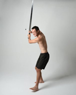 Yakışıklı Asyalı erkek modelin tam boy portresi, spor şortu ve gömleksiz. Beyaz stüdyo arka planında izole edilmiş, savaşçı eğitimi sırasında kılıçlarını tutuyor..