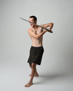 Yakışıklı Asyalı erkek modelin tam boy portresi, spor şortu ve gömleksiz. Beyaz stüdyo arka planında izole edilmiş, savaşçı eğitimi sırasında kılıçlarını tutuyor..