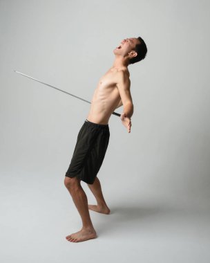 Yakışıklı Asyalı erkek modelin tam boy portresi, spor şortu ve gömleksiz. Beyaz stüdyo arka planında izole edilmiş, savaşçı eğitimi sırasında kılıçlarını tutuyor..