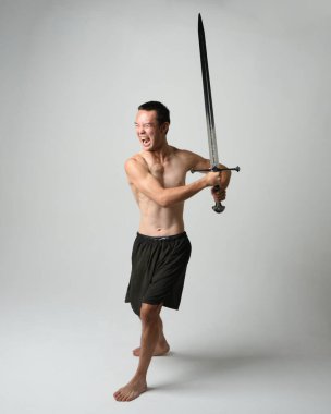 Yakışıklı Asyalı erkek modelin tam boy portresi, spor şortu ve gömleksiz. Beyaz stüdyo arka planında izole edilmiş, savaşçı eğitimi sırasında kılıçlarını tutuyor..