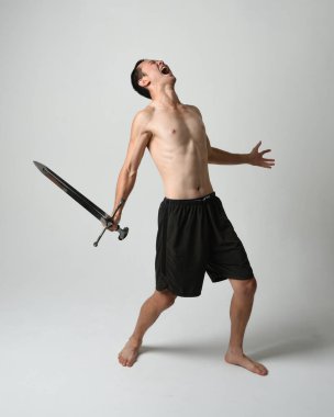 Yakışıklı Asyalı erkek modelin tam boy portresi, spor şortu ve gömleksiz. Beyaz stüdyo arka planında izole edilmiş, savaşçı eğitimi sırasında kılıçlarını tutuyor..