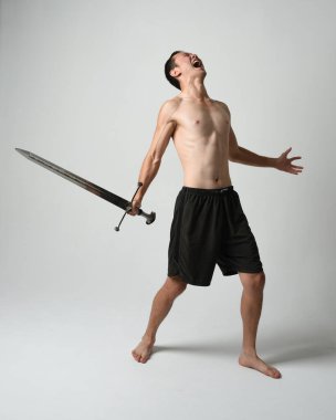Yakışıklı Asyalı erkek modelin tam boy portresi, spor şortu ve gömleksiz. Beyaz stüdyo arka planında izole edilmiş, savaşçı eğitimi sırasında kılıçlarını tutuyor..