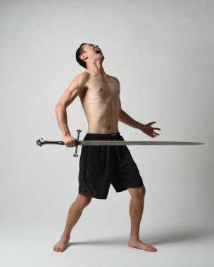 Yakışıklı Asyalı erkek modelin tam boy portresi, spor şortu ve gömleksiz. Beyaz stüdyo arka planında izole edilmiş, savaşçı eğitimi sırasında kılıçlarını tutuyor..