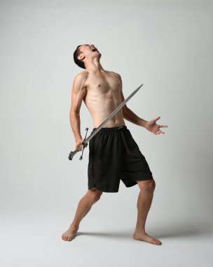 Yakışıklı Asyalı erkek modelin tam boy portresi, spor şortu ve gömleksiz. Beyaz stüdyo arka planında izole edilmiş, savaşçı eğitimi sırasında kılıçlarını tutuyor..