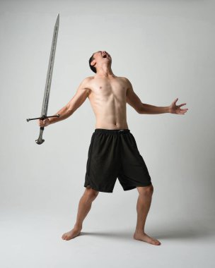 Yakışıklı Asyalı erkek modelin tam boy portresi, spor şortu ve gömleksiz. Beyaz stüdyo arka planında izole edilmiş, savaşçı eğitimi sırasında kılıçlarını tutuyor..