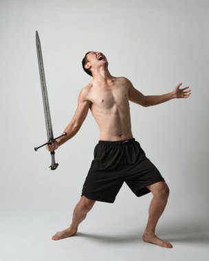 Yakışıklı Asyalı erkek modelin tam boy portresi, spor şortu ve gömleksiz. Beyaz stüdyo arka planında izole edilmiş, savaşçı eğitimi sırasında kılıçlarını tutuyor..