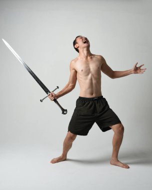Yakışıklı Asyalı erkek modelin tam boy portresi, spor şortu ve gömleksiz. Beyaz stüdyo arka planında izole edilmiş, savaşçı eğitimi sırasında kılıçlarını tutuyor..