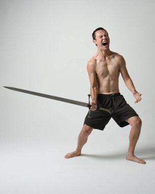 Yakışıklı Asyalı erkek modelin tam boy portresi, spor şortu ve gömleksiz. Beyaz stüdyo arka planında izole edilmiş, savaşçı eğitimi sırasında kılıçlarını tutuyor..