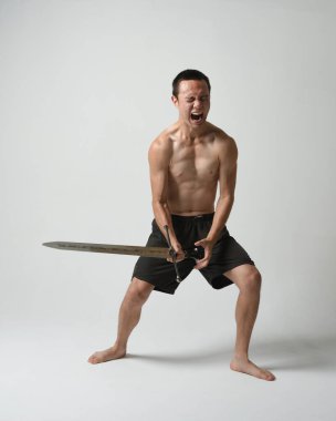 Yakışıklı Asyalı erkek modelin tam boy portresi, spor şortu ve gömleksiz. Beyaz stüdyo arka planında izole edilmiş, savaşçı eğitimi sırasında kılıçlarını tutuyor..