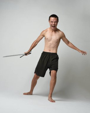 Yakışıklı Asyalı erkek modelin tam boy portresi, spor şortu ve gömleksiz. Beyaz stüdyo arka planında izole edilmiş, savaşçı eğitimi sırasında kılıçlarını tutuyor..