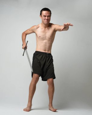 Yakışıklı Asyalı erkek modelin tam boy portresi, spor şortu ve gömleksiz. Beyaz stüdyo arka planında izole edilmiş, savaşçı eğitimi sırasında kılıçlarını tutuyor..