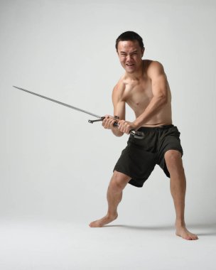 Yakışıklı Asyalı erkek modelin tam boy portresi, spor şortu ve gömleksiz. Beyaz stüdyo arka planında izole edilmiş, savaşçı eğitimi sırasında kılıçlarını tutuyor..