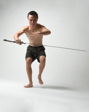 Yakışıklı Asyalı erkek modelin tam boy portresi, spor şortu ve gömleksiz. Beyaz stüdyo arka planında izole edilmiş, savaşçı eğitimi sırasında kılıçlarını tutuyor..