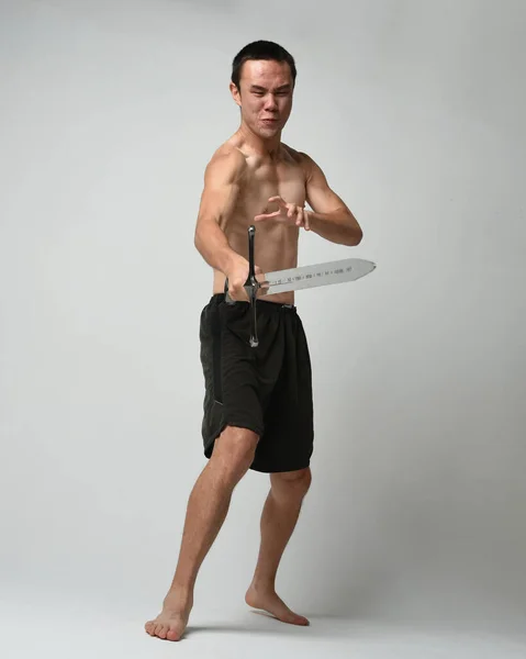 Full length portrait of fit handsome asian male model, wearing gym shorts and shirtless. Holding sword weapons  whilst in warrior training action poses, isolated on white studio background.