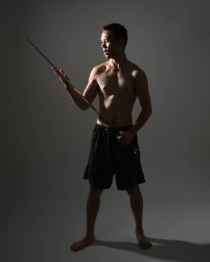 Yakışıklı Asyalı erkek modelin portresi, spor şortu ve üstsüz. Savaşçı eğitimleri sırasında kılıçlarını tutarken kasvetli karanlık stüdyo arka planında siluet ile izole edilmiş pozlar veriyordu.