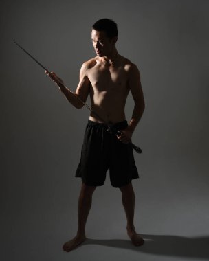 Yakışıklı Asyalı erkek modelin portresi, spor şortu ve üstsüz. Savaşçı eğitimleri sırasında kılıçlarını tutarken kasvetli karanlık stüdyo arka planında siluet ile izole edilmiş pozlar veriyordu.