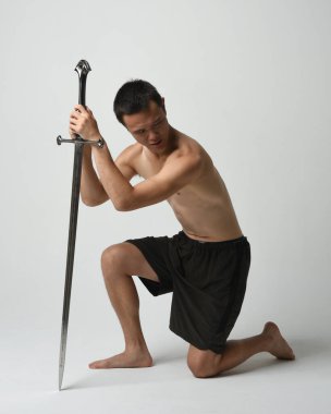 Yakışıklı Asyalı erkek modelin tam boy portresi, spor şortu ve gömleksiz. Kılıç tutan, savaşçı eğitim pozisyonunda diz çöken, beyaz stüdyo arka planında izole edilmiş..