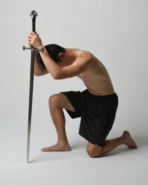 Yakışıklı Asyalı erkek modelin tam boy portresi, spor şortu ve gömleksiz. Kılıç tutan, savaşçı eğitim pozisyonunda diz çöken, beyaz stüdyo arka planında izole edilmiş..