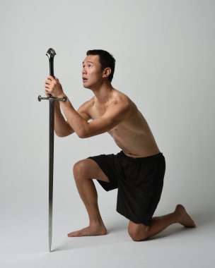 Yakışıklı Asyalı erkek modelin tam boy portresi, spor şortu ve gömleksiz. Kılıç tutan, savaşçı eğitim pozisyonunda diz çöken, beyaz stüdyo arka planında izole edilmiş..